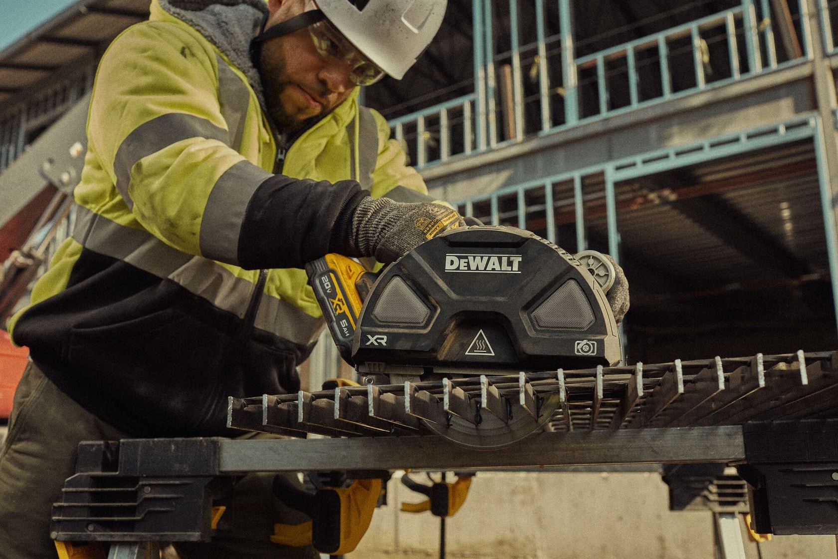 A construction worker using a DEWALT DCS383H1 cordless saw to cut a metal grate at a construction site. The worker is wearing a yellow and black safety jacket, gloves, and a helmet. The saw is positioned on the metal surface, and there is a partially built structure in the background.