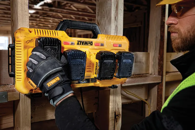Eine Person mit Arbeitshandschuhen installiert oder entfernt Batterien aus einem gelben DEWALT Multi-Batterieladegerät, das an einem Holzbalken auf einer Baustelle befestigt ist.