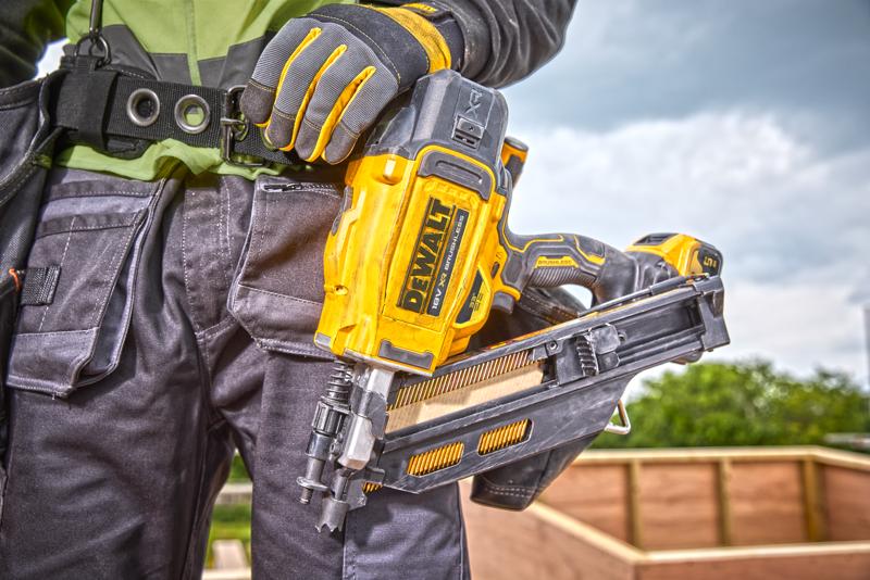 Person mit DEWALT DCN930N Nagelpistole, Handschuhen und Arbeitskleidung an einer Baustelle im Freien.