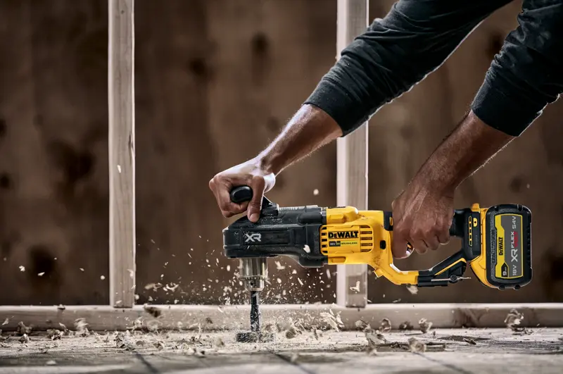 Eine Person benutzt einen DEWALT DCD444N Akku-Bohrmaschine mit Seitengriff, um durch Holz zu bohren. Dabei entstehen Holzspäne und Staub. Im Hintergrund ist eine Baukonstruktion zu sehen.