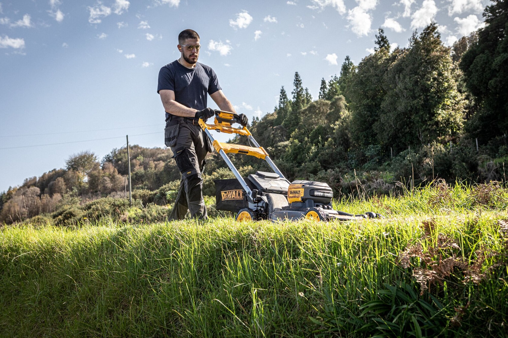 Person mäht draußen Gras mit einem DEWALT Rasenmäher, sonniger Tag, Bäume und blauer Himmel im Hintergrund.