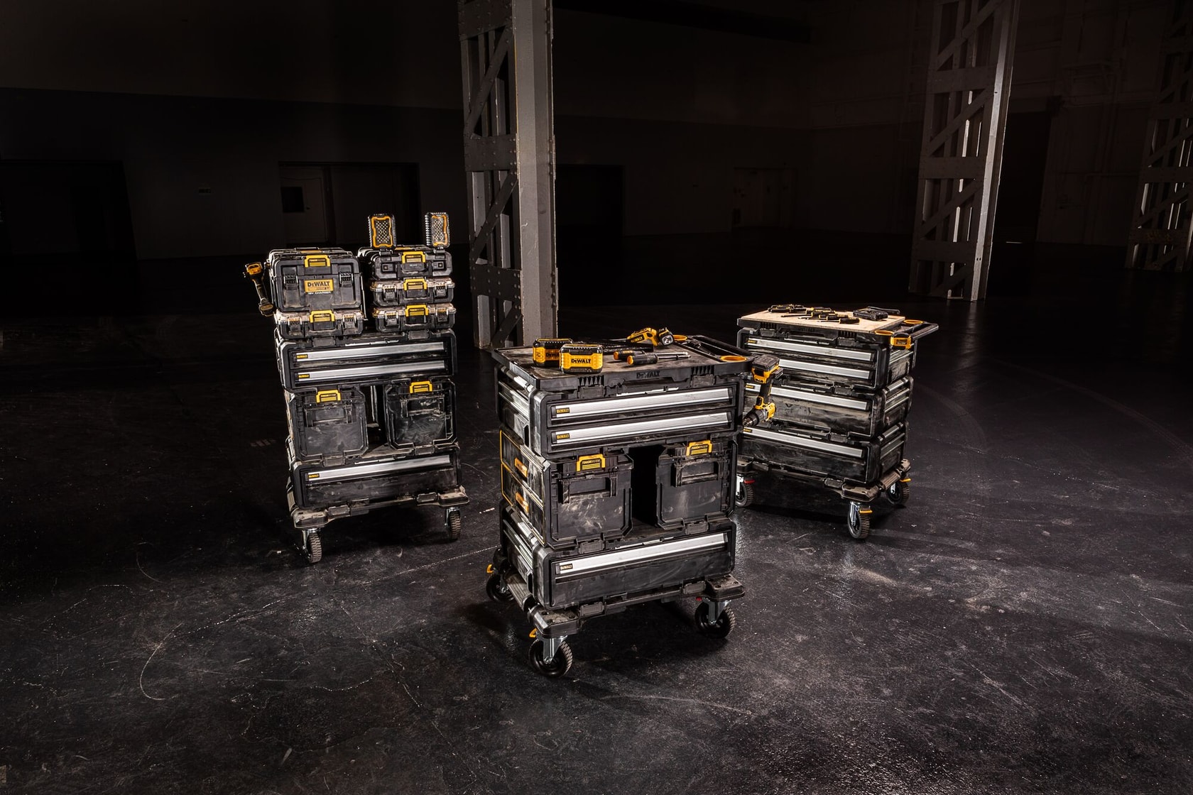 Drei fahrbare Stapel DEWALT-Werkzeugkisten mit Rädern stehen in einem dunklen Industrieumfeld. Jeder Stapel besteht aus mehreren schwarzen und gelben Aufbewahrungsboxen zur Organisation und zum Transport von Werkzeugen.